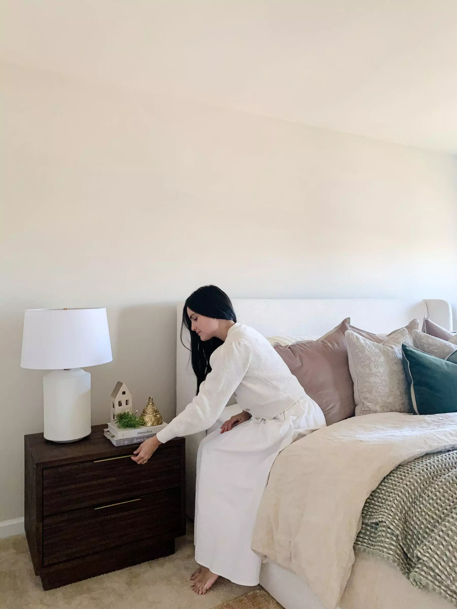 A woman is sitting on her bed as she reaches into her wooden nightstand.