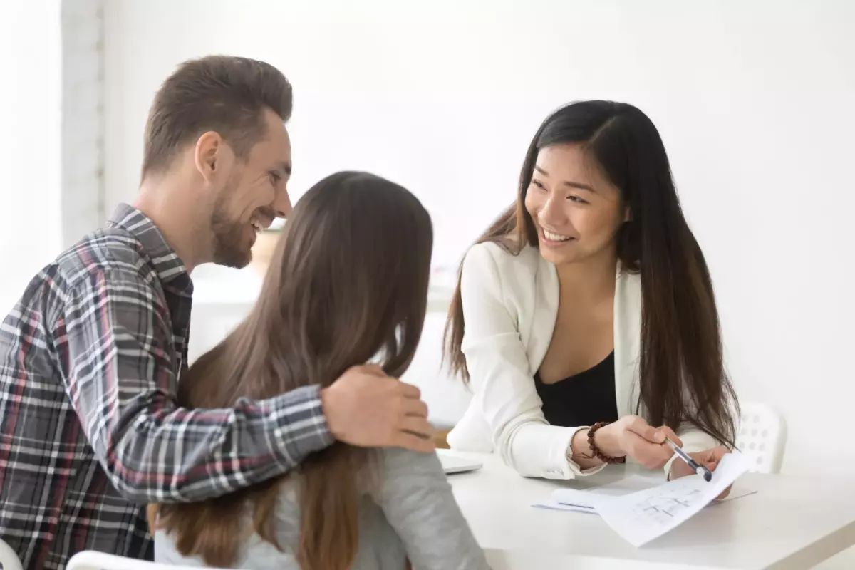 A man and woman with their backs to the camera sit across a table from a woman showing them paperwork for forming an LLC for real estate investments.