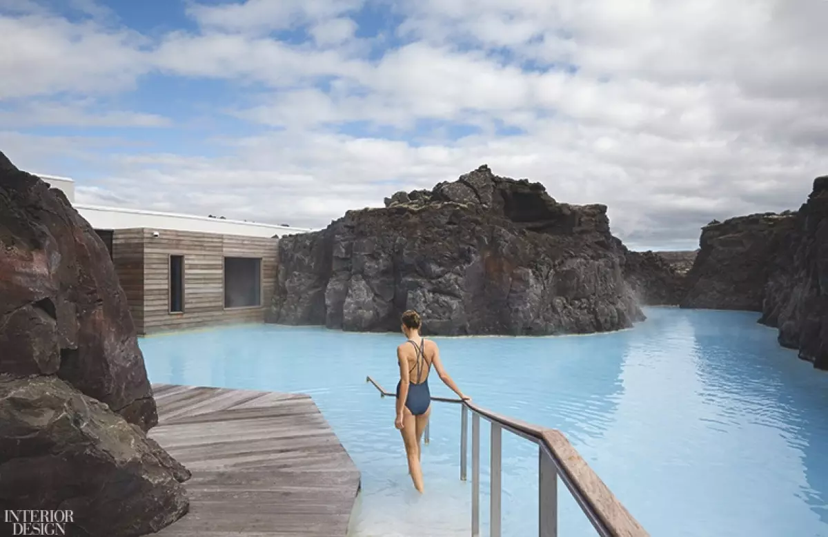 A woman steps into a blue lagoon surrounded by rock formations while holding a wooden railing