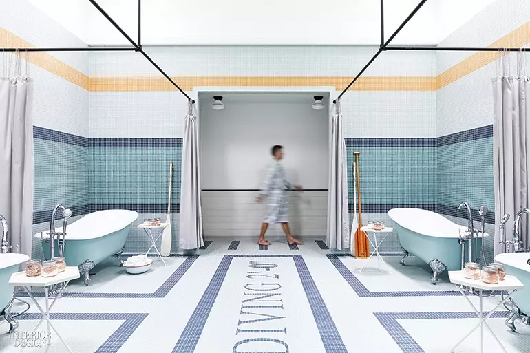 A man walks in a room lined with blue tile and a pair of light blue clawfoot bathtubs