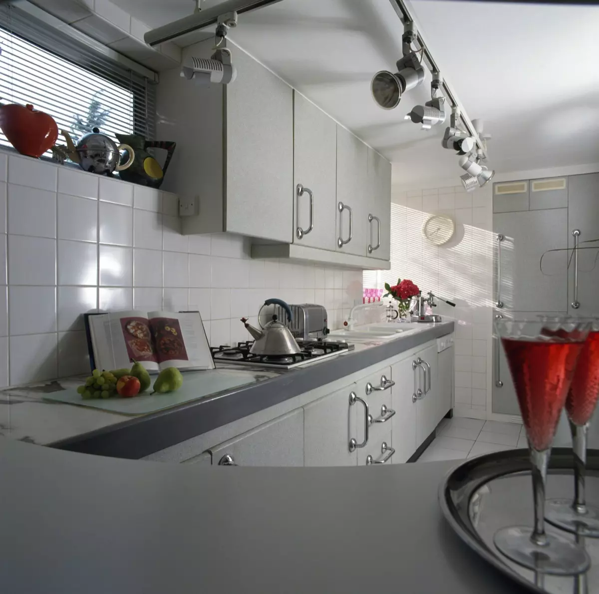 Track spotlights and white tiling in a nineties kitchen with pale gray fitted units and an Alessi kettle on the hob