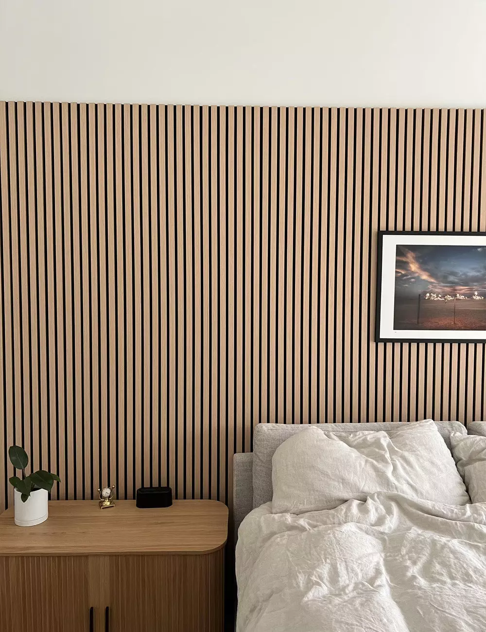 White bedroom with an indoor olive tree, framed art, and a slatted wood headboard