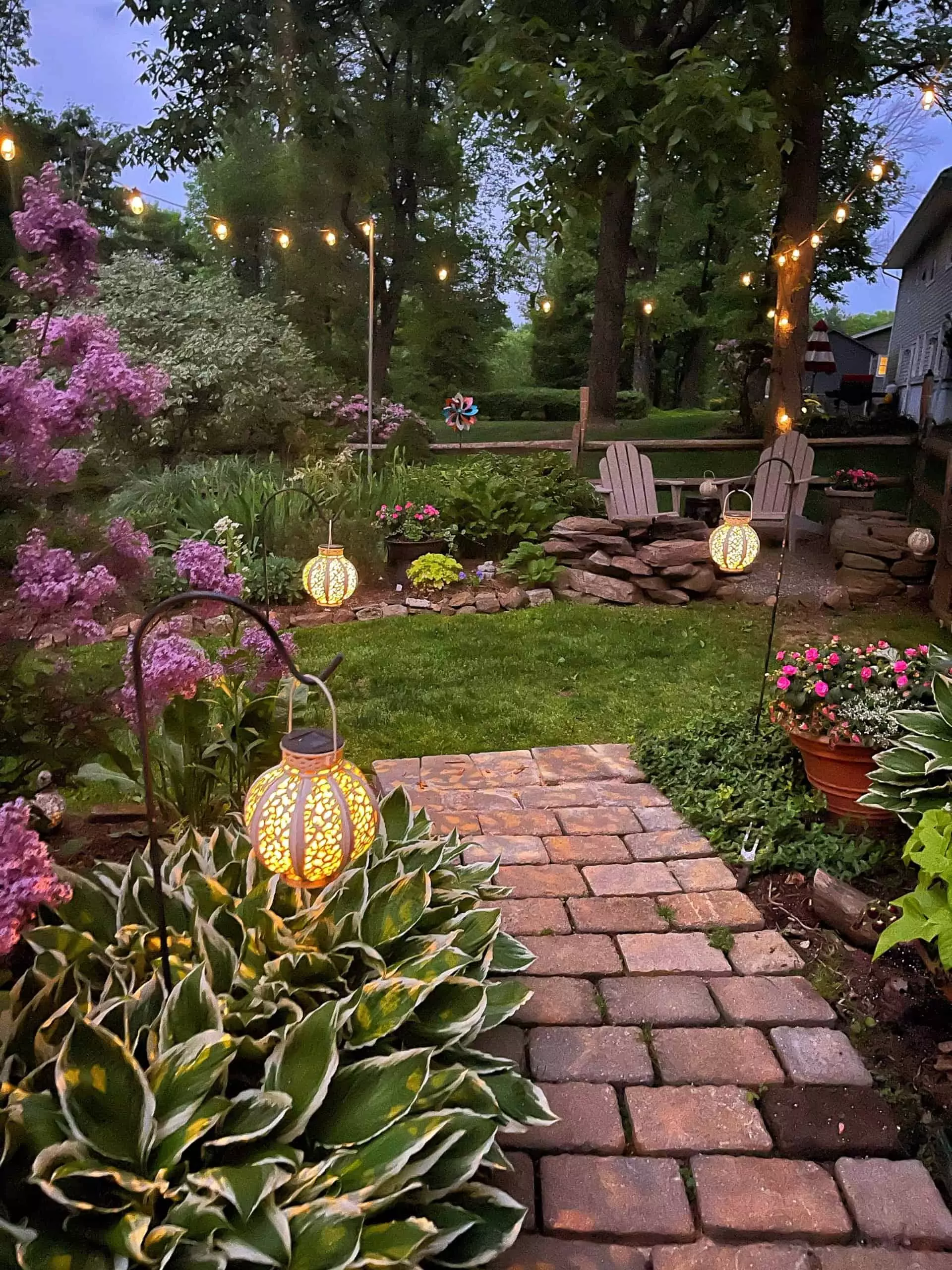 Lilac flowers by the Fire Pit Garden at night lit with solar lanterns and surrounded by hostas and twinkle lights