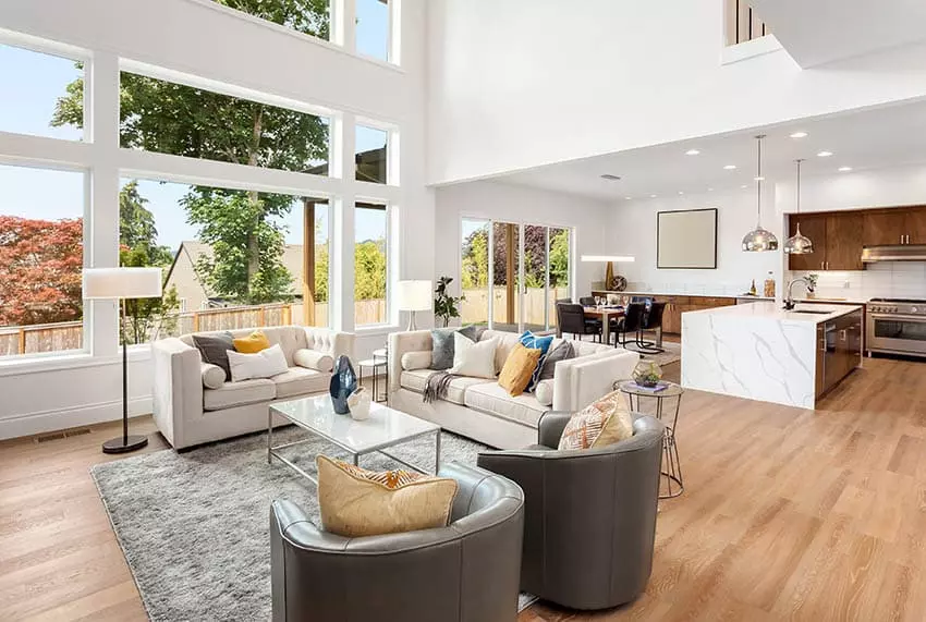 Living room with transitional sofa next to a kitchen with white cabinets, island, high chairs, backsplash, and wood flooring