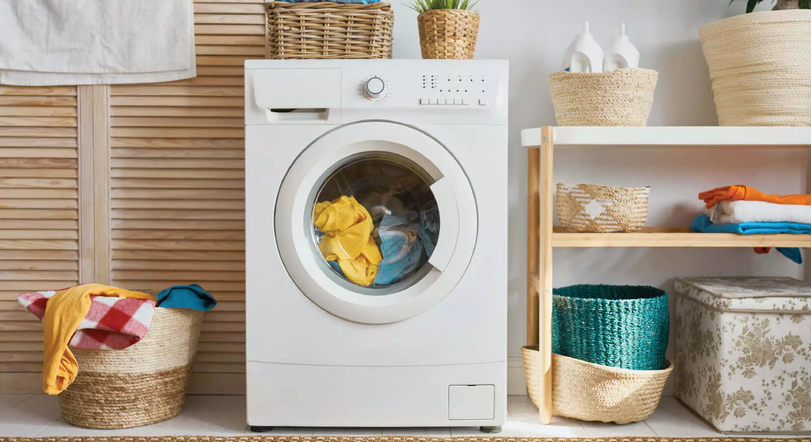 Washer with a load of laundry next to a hamper of laundry and a wood louvered-style cabinet.