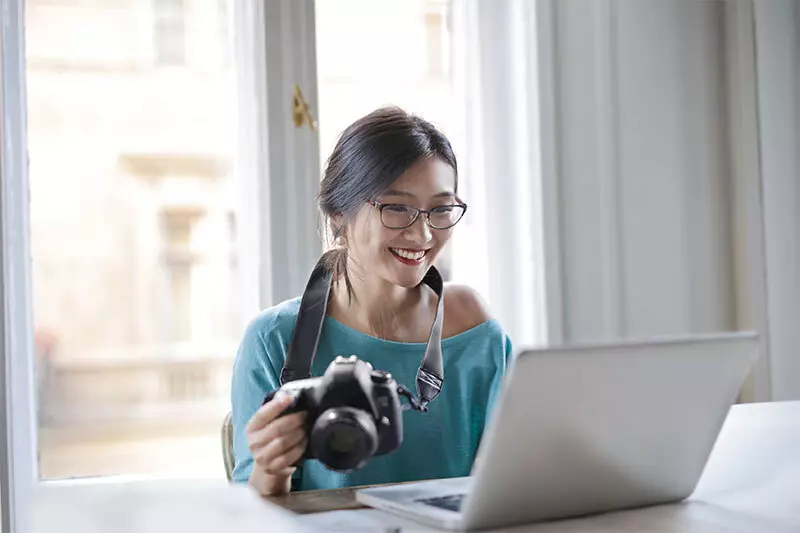 Cheerful woman with camera using laptop