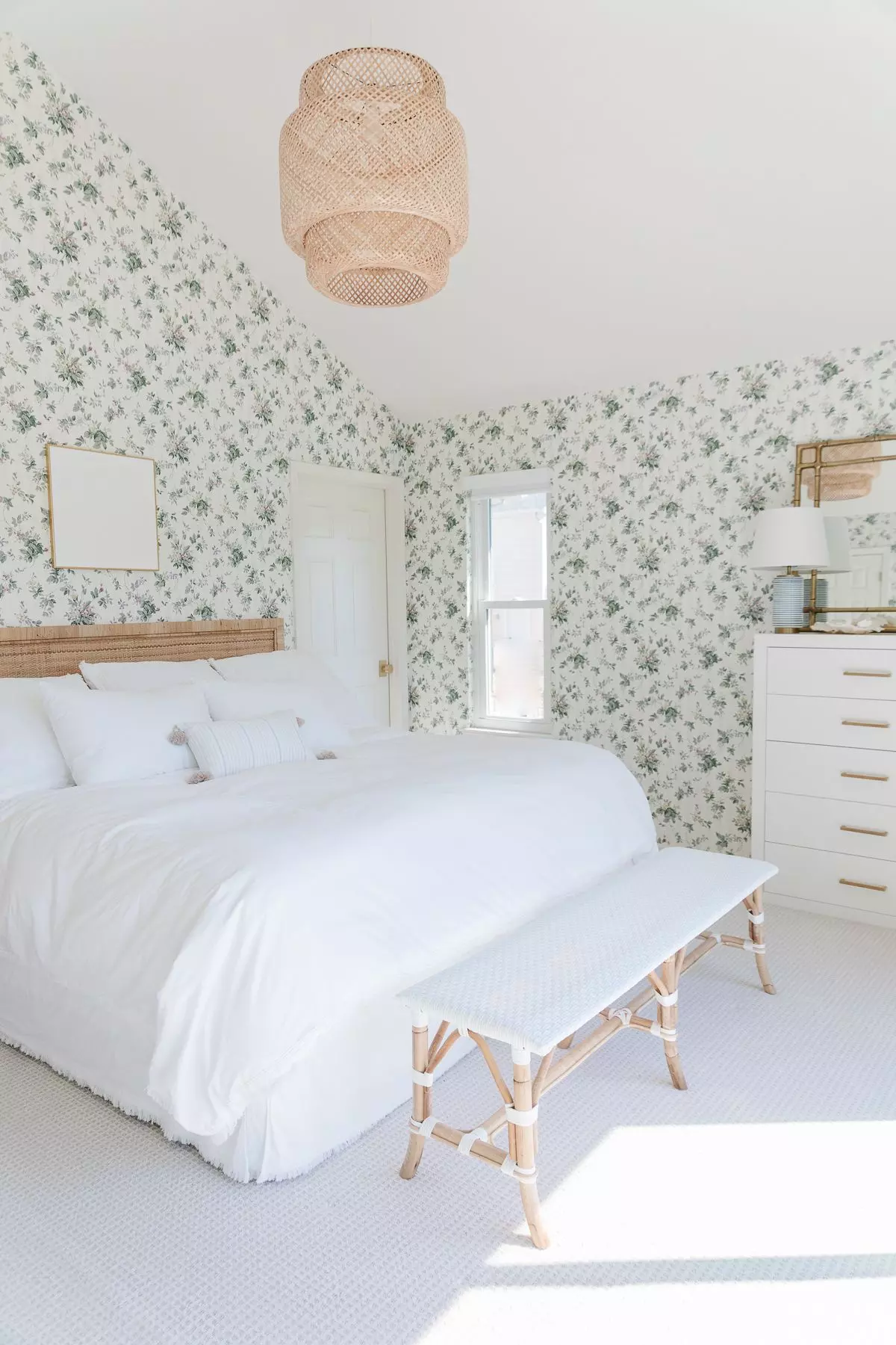 A coastal bedroom with floral wallpaper, a rattan headboard, bench, and light fixture