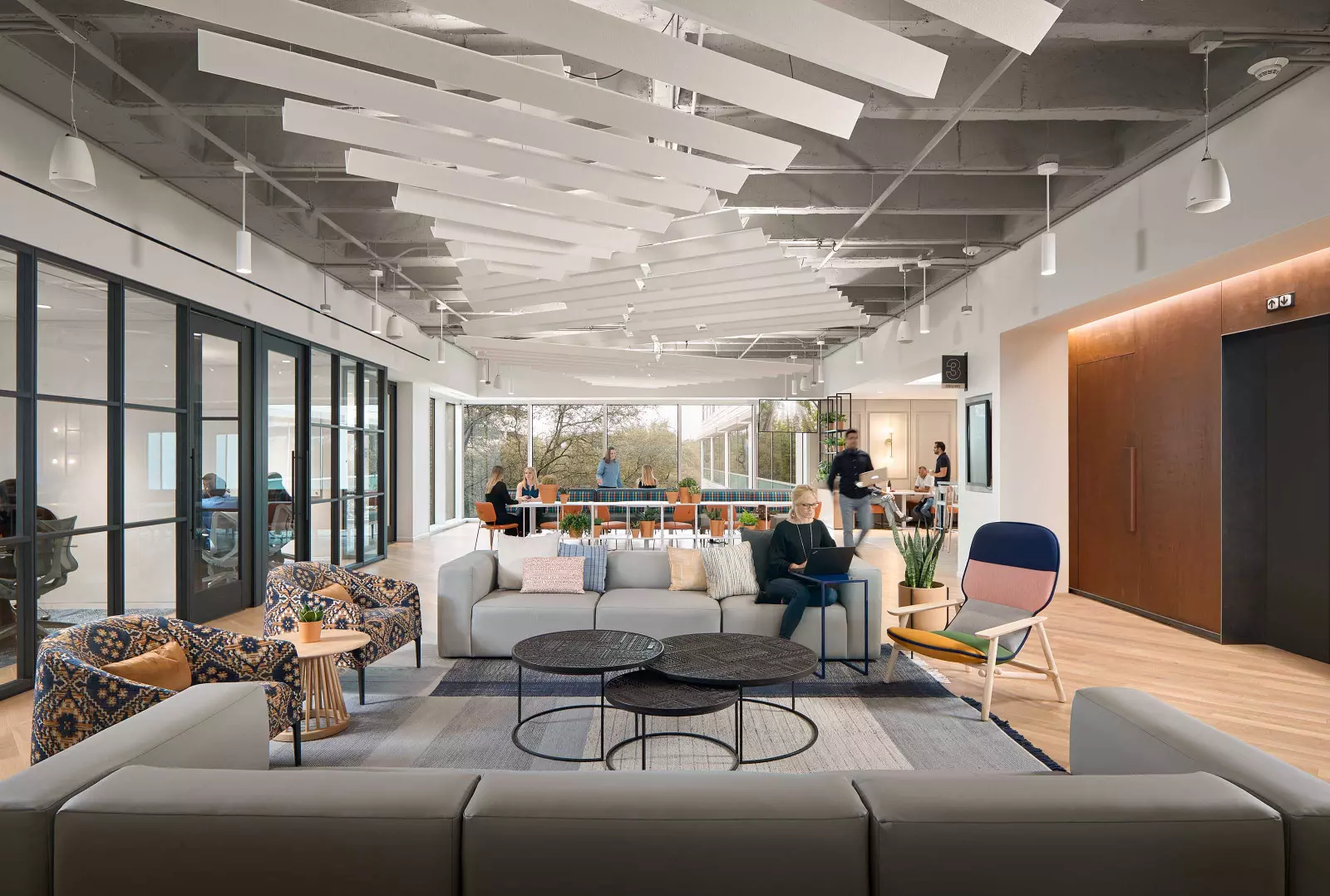 This seating area features two gray sofas, three patterned chairs, three coffee tables, and pillows of various colors. There is a person sitting on one of the sofas working on their computer.