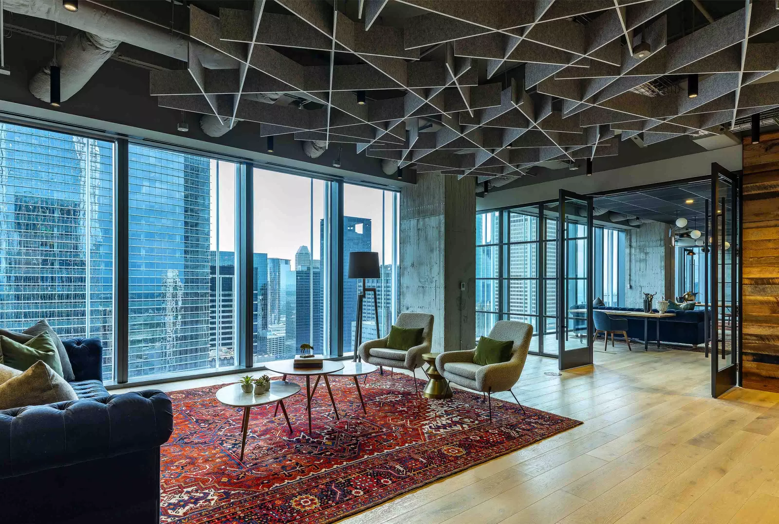 This lounge area has a red patterned rug, two green chairs, three coffee tables, and a blue sofa. It has large windows that overlook the city.