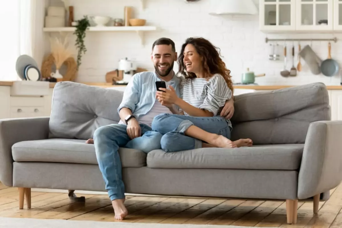 Happy young woman and man hugging, using smartphone together, sitting on cozy couch at home