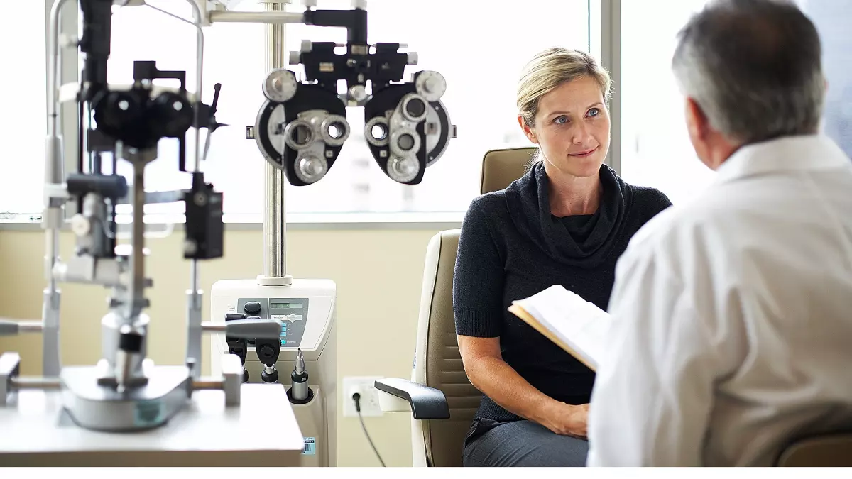 A woman is at an eye exam with her doctor. They’re off to the right of the frame, and the left of the frame is the eye exam equipment.