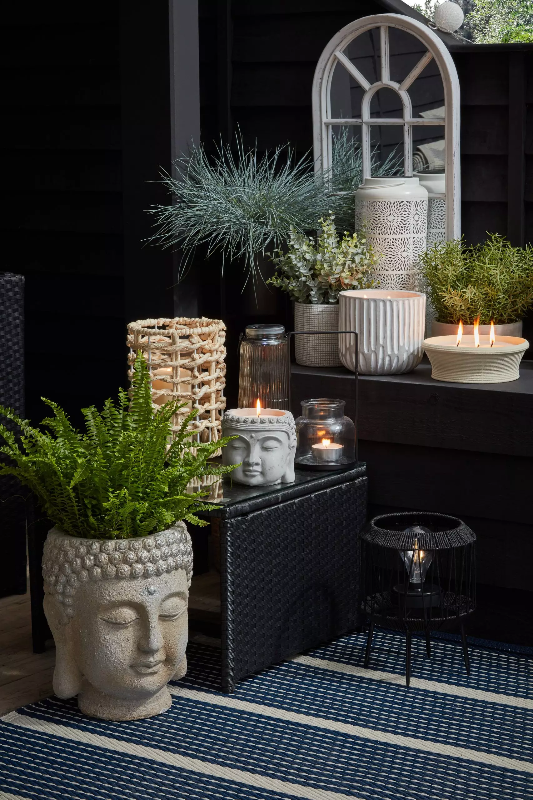 Black corner of a garden with buddha head plant pots and candles, rattan and concrete candle holders, and an arched outdoor mirror
