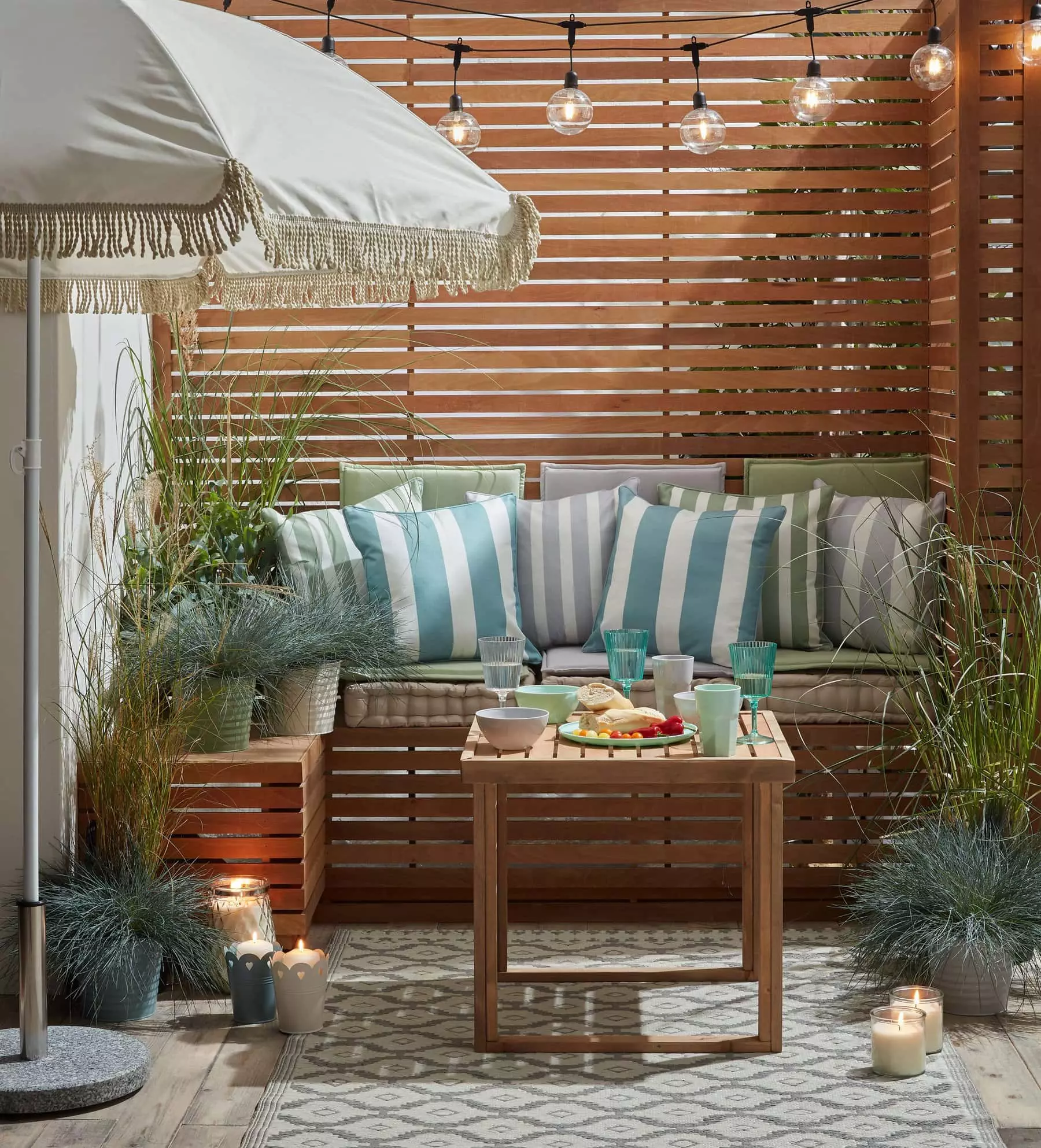 Panelled fence with striped cushions, pallet furniture, outdoor rug, and tasseled parasol in the garden