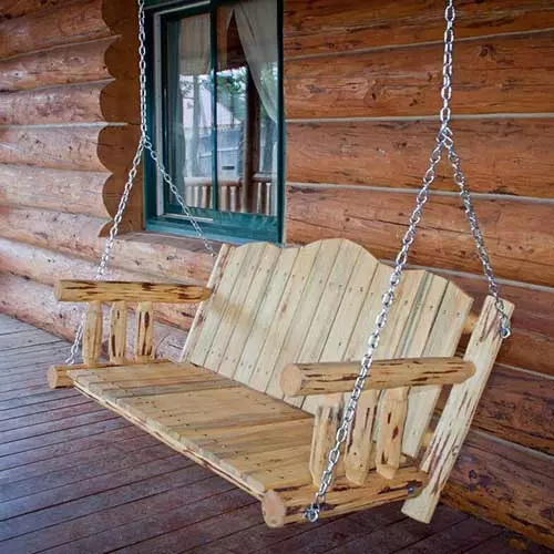 A wicker hanging bench seat with rounded edges and a dark blue cushion on a deck, with the side of a house and garden scene in the background in soft focus.