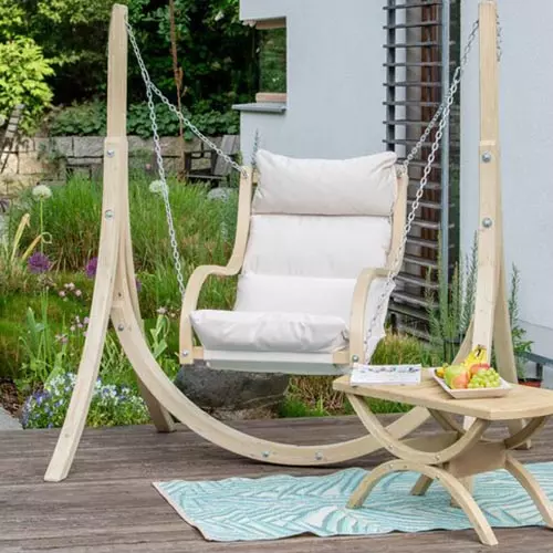 A circular hanging seat, in black wicker with white striped cushions hanging over a concrete patio with a swimming pool and the ocean in the background.