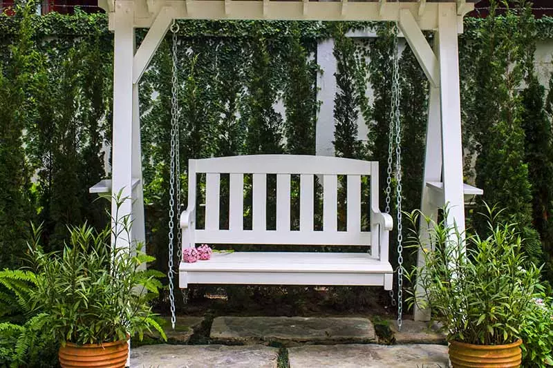 A close-up of a wooden hanging bench seat with scatter cushions and a striped throw rug, set on a wooden deck with a garden scene in the background.