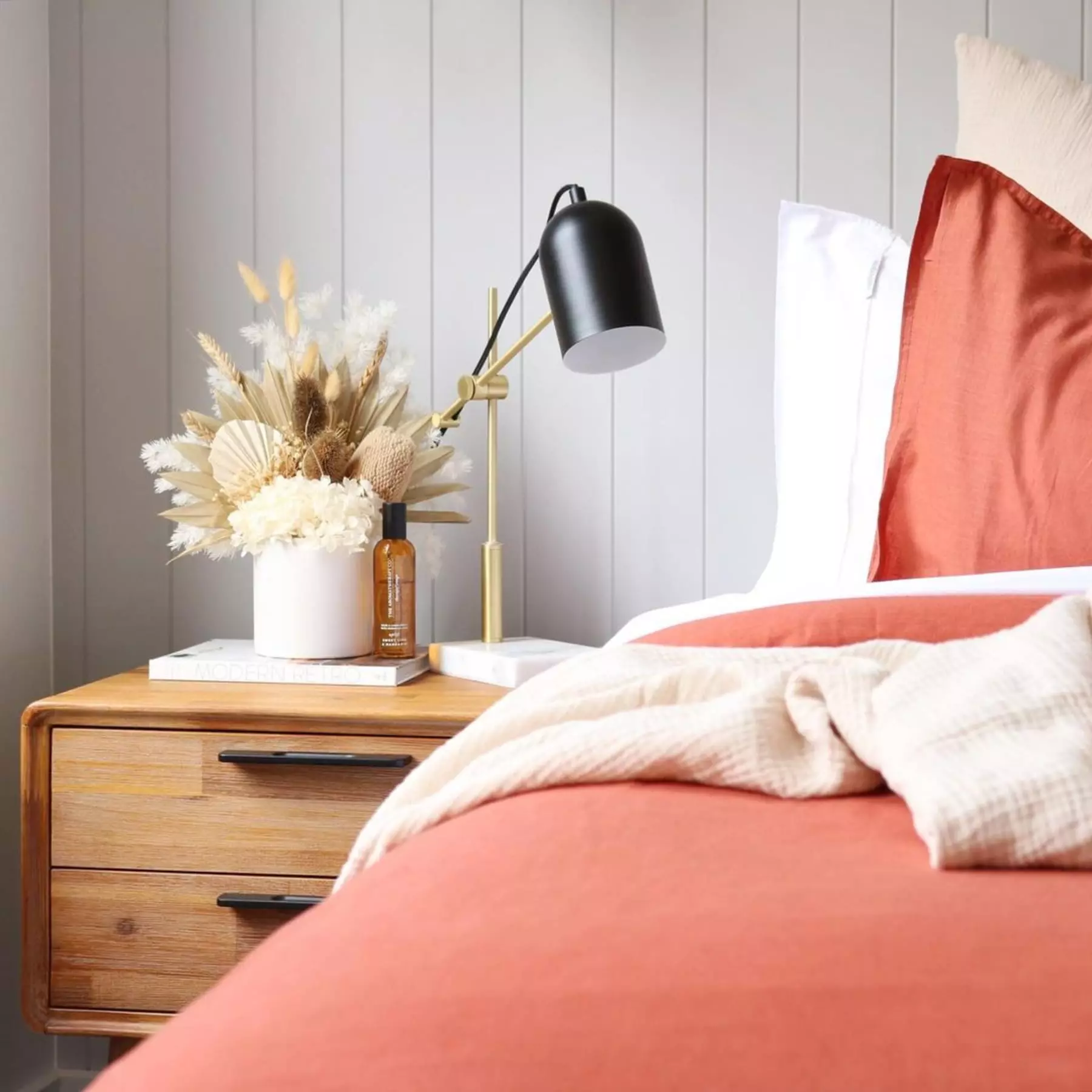 A bed in a bright coral sheet and a wooden bedside table decorated with a lamp and a vase.