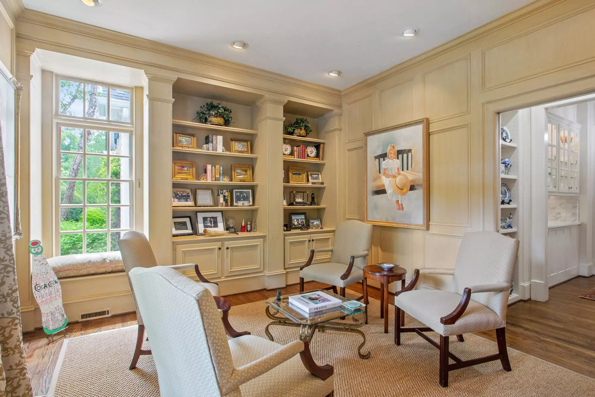 Whitewashed light-colored antique wood walls in a beautiful sitting room. Built-in shelving and cabinetry, wood floors and a beige area rug. Dark wood chairs with light-colored cream cushions. Window seat.