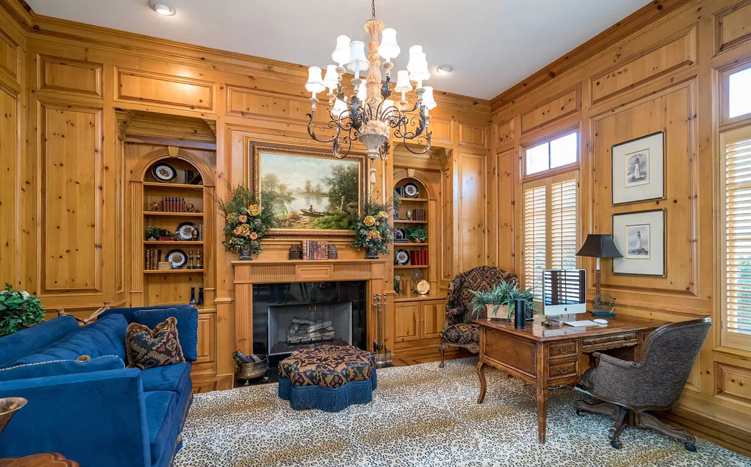 Home office with all wood walls and light brown stain. This stain is so light it's actually just a seal. Knotty pine wall panels. Gas fireplace with black surround and wood mantle. Built-ins. Matching hardwood floors with area rug. Plush blue sofa. Wood desk. White sheetrock ceiling with center chandelier.