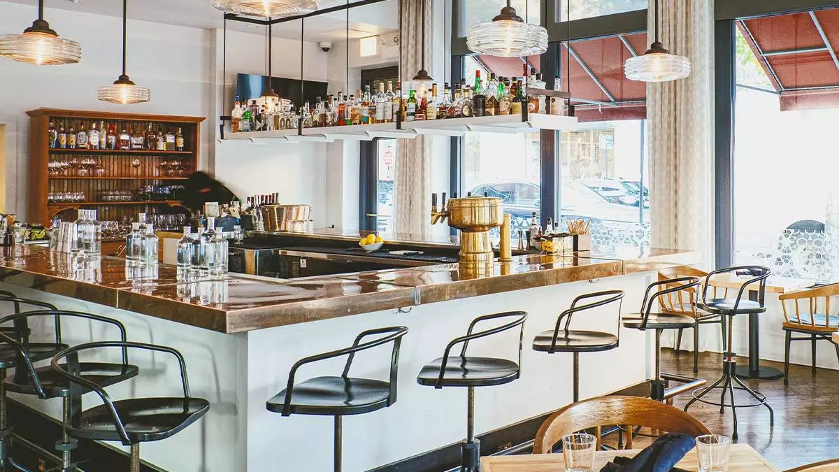 A dining area with grey chairs and wooden tables with lots of greenery surrounding it and a bar in the background with large light fixtures and a wall of bottles at Aba Restaurant in Chicago, Illinois, USA