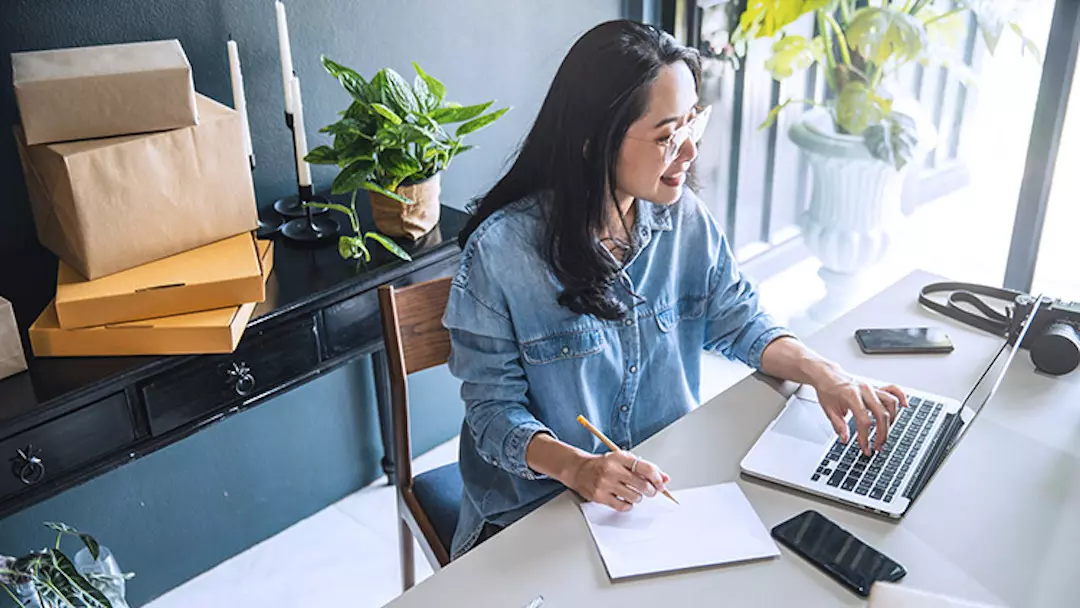 A woman types on her laptop while taking notes in her home office